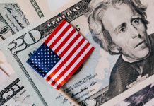 Fed Chairman Warsh on Global Policy, Declines Rate Guidance Overhead view of a small U.S. flag atop a stack of $20 bills, symbolizing national business and finance.